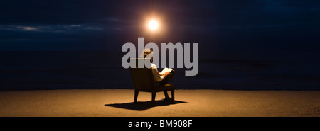 Man sitting in chair sous la lumière ampoule sur plage le soir, reading book Banque D'Images