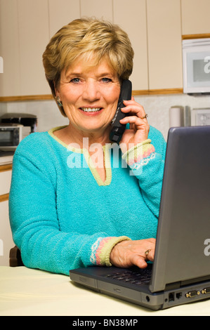 Travail à domicile, a senior woman multitâches : Prend un appel téléphonique tout en travaillant sur un ordinateur portable, tous à sa table de cuisine. Banque D'Images