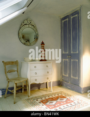 Miroir vénitien mural au-dessus de petite chaise et poitrine blanche-de-tiroirs dans une chambre mansardée avec effet en détresse porte peinte en bleu Banque D'Images