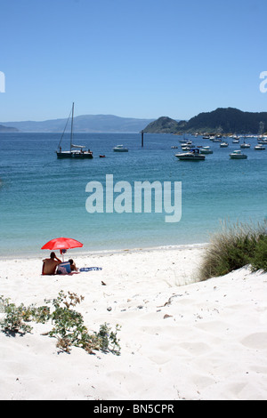 Plage de Praia de Rodas, avec couple sous red parasol et de bateaux dans la baie, l'Illa de Faro, Illas Cies, Vigo, Galice, Espagne du Nord Banque D'Images