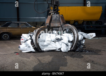 Voiture écrasée cube dans les mâchoires d'une grue mécanique en face de l'aplatisseur dans un parc à ferrailles - maintenant appelé le recyclage du véhicule Banque D'Images