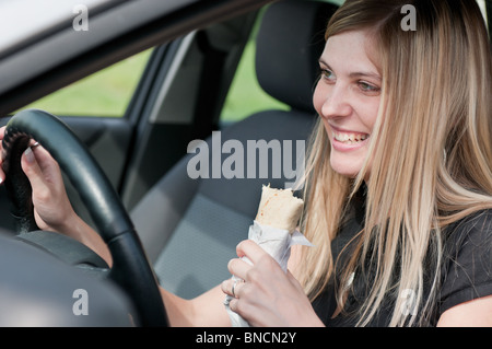 Young beautiful smiling woman driving car and eating fast food - portrait par fenêtre latérale Banque D'Images