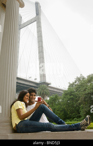 Couple looking at mobile phone avec le pont en arrière-plan, James Famille Prinsep Memorial, Vidyasagar Setu, Kolkata, West Bengal, India Banque D'Images