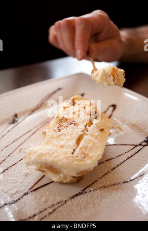 Une personne dégustant une tranche de gâteau Napoléon crémeux, garnie de sucre en poudre et de sirop de chocolat, sur une plaque de céramique blanche. Banque D'Images