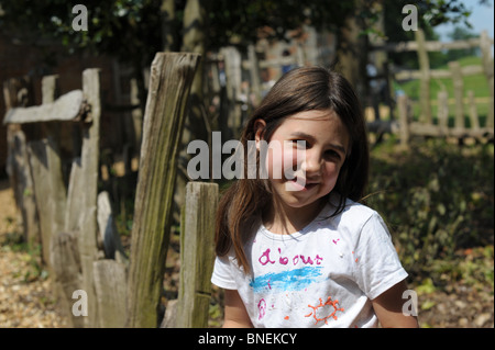 Petite fille aux cheveux long noir assis dans un parc à l'été de porter un T shirt blanc avec un soleil sur elle. Elle sourit. Banque D'Images