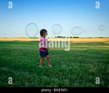 Jeune fille hispanique s'exécute dans l'herbe verte de l'été qu'elle a le pouvoir d'énormes bulles de savon dans la vaste étendue d'un crépuscule à mi-américain Banque D'Images