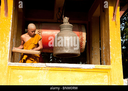 Un moine bouddhiste novice sonne la cloche de bronze avec un morceau de bois à son temple de Luang Prabang Laos Banque D'Images