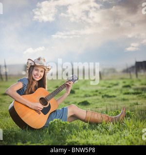 Cowgirl sitting in field playing guitar Banque D'Images