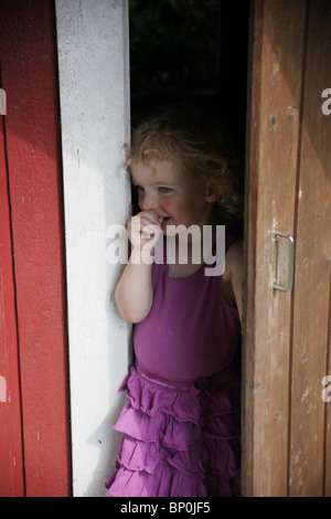 Petite fille vêtements enfant en partie de sucer son pouce à l'extérieur de l'ouverture d'un modèle de porte en bois playhouse publié Banque D'Images