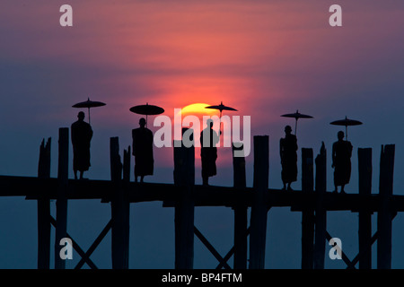 Les moines d'Asie se tenir sur le pont en teck U Bein dans Amarapura, Myanmar. Le soleil couchant est suspendu au-dessus de leurs silhouettes. Banque D'Images