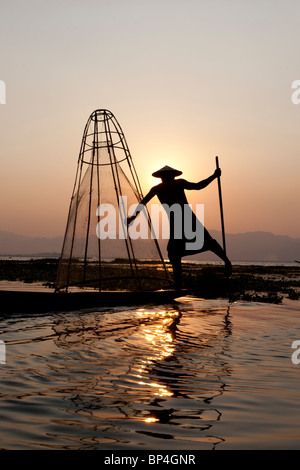 Un pêcheur d'aviron de la jambe de bois de teck ses manœuvres canoe au-dessus du poisson sur le lac alors que le soleil se couche derrière lui exposant son corps un Banque D'Images