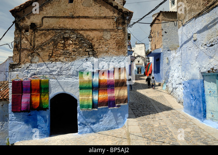 Scène de rue à Chefchaouen avec affichage de tapis marocain à vendre accroché sur les murs, & Blue, maisons Maroc Banque D'Images