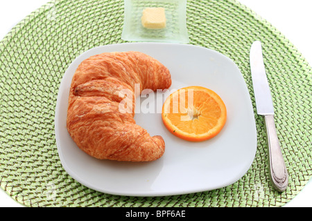Le croissant d'or à tranche d'orange sur une assiette blanche avec un couteau et une pointe de beurre sur un napperon en rotin vert Banque D'Images