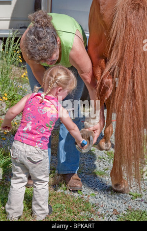 Cette grand-mère aide son enfant en petite-fille blonde avec des nattes tressées pour aider à nettoyer le sabot du cheval en préparation. Banque D'Images