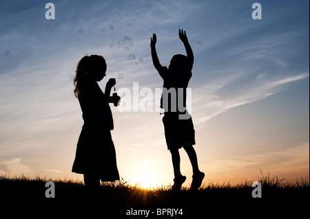 Silhouette d'une jeune fille et garçon soufflant et jouant avec des bulles au coucher du soleil. Banque D'Images