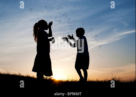 Silhouette d'une jeune fille et garçon soufflant et jouant avec des bulles au coucher du soleil. Banque D'Images