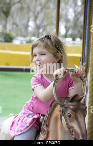 Funny happy gesturing petite blonde fille jouant sur les chevaux merry go round Banque D'Images