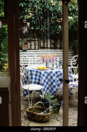 Vue à travers des portes françaises de table avec un chiffon blanc et bleu controle metal chaises dans le patio du gravier dans petite ville jardin Banque D'Images