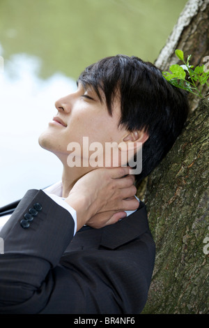 Young businessman leaning against tree trunk, profile Banque D'Images