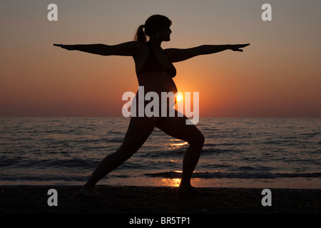 Une femme enceinte pratique le yoga sur une plage de Toscane, silhoutted par le soleil couchant. Banque D'Images