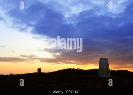 Trig point et pierre commémorative en silhouette contre le ciel coloré au lever du soleil sur Lealholm Moor près de Danby Beacon Banque D'Images