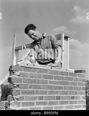 1940 HOMME TRAVAILLANT AVEC NIVEAU SUR MUR DE BRIQUE DE CONSTRUCTION DE CHEMINÉE Banque D'Images