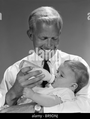 1960 SMILING BLONDE PÈRE ENFANT ALIMENTATION DE LAIT DANS LE BIBERON EN VERRE Banque D'Images