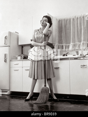 1950 FEMME AU FOYER FEMME DEBOUT DANS LA CUISINE S'APPUYANT SUR LE BALAI Banque D'Images