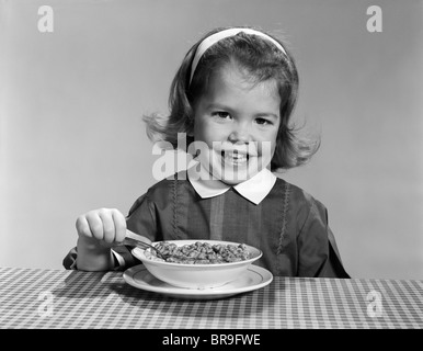 Années 1950 Années 1960 SMILING LITTLE GIRL EATING UN BOL DE CÉRÉALES POUR PETIT DÉJEUNER LOOKING AT CAMERA Banque D'Images