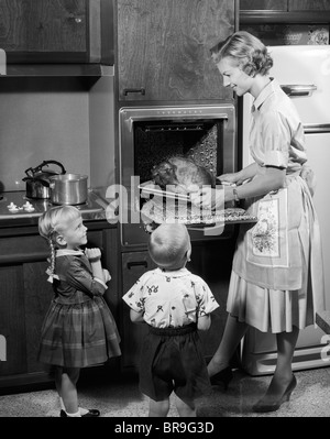 Années 1950, FILLE ET FILS REGARDANT MÈRE déposer le rôti de dinde de Thanksgiving LE DÎNER DE CUISINE Four électrique Banque D'Images