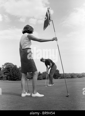 1970 COUPLE PLAYING GOLF MAN PUTTING WOMAN HOLDING FLAG Banque D'Images