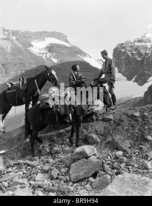 Années 1920 Années 1930 COUPLE HOMME FEMME PORTANT DES VÊTEMENTS D'ÉQUITATION ÉPERONS BOTTES ASSIS DEBOUT SUR GROS ROCHER PAR DEUX CHEVAUX Banque D'Images