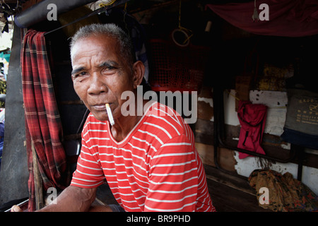 Un homme bénéficie d'une cigarette tout en étant assis sur sa maison dans un village de squatteurs à Phnom Penh, Cambodge. Banque D'Images