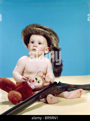 1960 BABY BOY WEARING COONSKIN CAP AVEC ANIMAL EN PELUCHE ET DE FUSIL DE GUN Banque D'Images