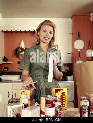 1960 JEUNE FEMME AU FOYER BLONDE SOURIANT LORS DE LA VÉRIFICATION DE LA RÉCEPTION D'ÉPICERIE DANS LA CUISINE Banque D'Images