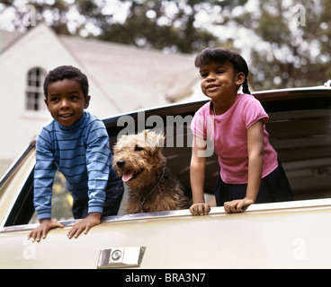 Des années 1970, peu d'Africains-Américains BOY AND GIRL LEANING OUT OF CAR FENÊTRE AVEC CHIEN AIREDALE HEUREUX vacances voyage Banque D'Images