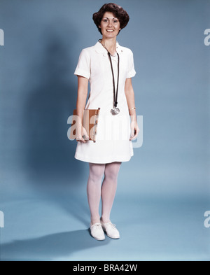 1970 COMITÉ PERMANENT FULL LENGTH PORTRAIT OF WOMAN SMILING NURSE WEARING WHITE UNIFORM AND STETHOSCOPE Banque D'Images