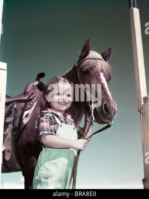 Années 1940 Années 1950 SMILING GIRL HOLDING PERMANENT SALOPETTE PAR CHEVAL RÊNES Banque D'Images