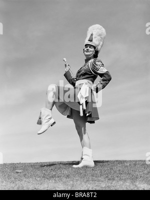 1950 SMILING majorette posant à l'EXTÉRIEUR EN UNIFORME AVEC JUPE COURTE bottes bottes hautes FURRY HAT tenant un baton Banque D'Images