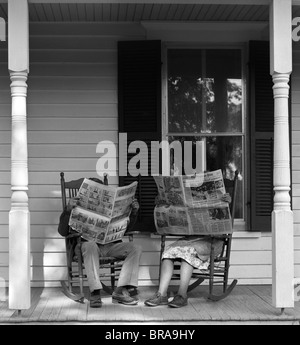 Années 1950 Années 1960 HOMME FEMME COUPLE SITTING ON PORCH DANS DES CHAISES À BASCULE HOLDING JOURNAUX PAR se cacher leur visage Banque D'Images