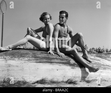 1930 HOMME FEMME COUPLE SMILING PORTANT DES MAILLOTS assis dos à dos SUR LE BATEAU EN BOIS RENVERSÉE Banque D'Images