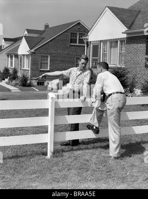 1950 2 HOMMES VOISINS DE CONVERSATIONS AU CLÔTURE EN BOIS BLANC ENTRE LES MAISONS DE BANLIEUE Banque D'Images