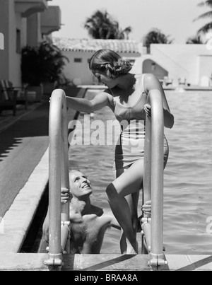 1930 FEMME PORTANT UNE PIÈCE MAILLOT ESCALADE DE PISCINE EN REGARDANT MAN SMILING Banque D'Images