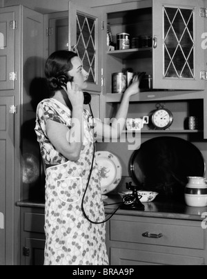 1930 FEMME AU FOYER FEMME PORTANT UN TABLIER DE CUISINE À METTRE DES ALIMENTS SUR MEUBLE étagère Banque D'Images