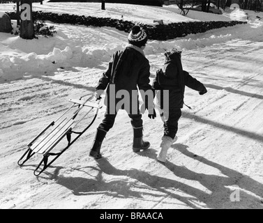 1970 VUE ARRIÈRE DE DEUX ENFANTS MARCHER DANS LA NEIGE TRAÎNEAU TIRANT Banque D'Images