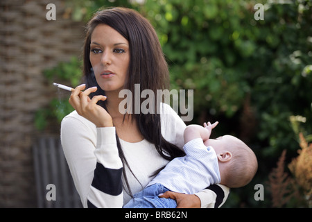 Mère dans sa vingtaine de fumer une cigarette et tenant son bébé dans les bras. Banque D'Images