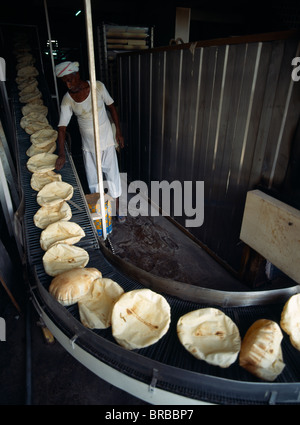 Doha Qatar Homme Baker dans une boulangerie à côté de la courroie du convoyeur transportant rond plat pains arabes sortant du four Banque D'Images