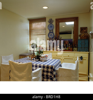 Meuble blanc-couvre sur des chaises à table avec un chiffon bleu vérifiée en petite salle à manger avec sol en bois store vénitien sur la fenêtre Banque D'Images