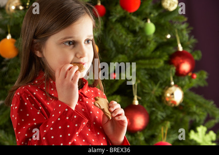 Young Girl Eating Star Shaped Christmas Cookie in front of Christmas Tree Banque D'Images