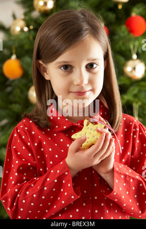 Young Girl Eating Star Shaped Christmas Cookie in front of Christmas Tree Banque D'Images